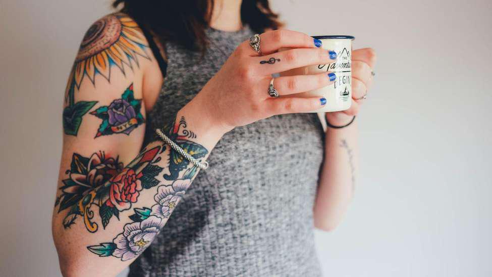 A woman holding a cup of tea. She is wearing a grey top and has colorful tattoos on her arm of various flowers, including a red rose and a sunflower. She has two rings on her fingers and a bracelet on her wrist.