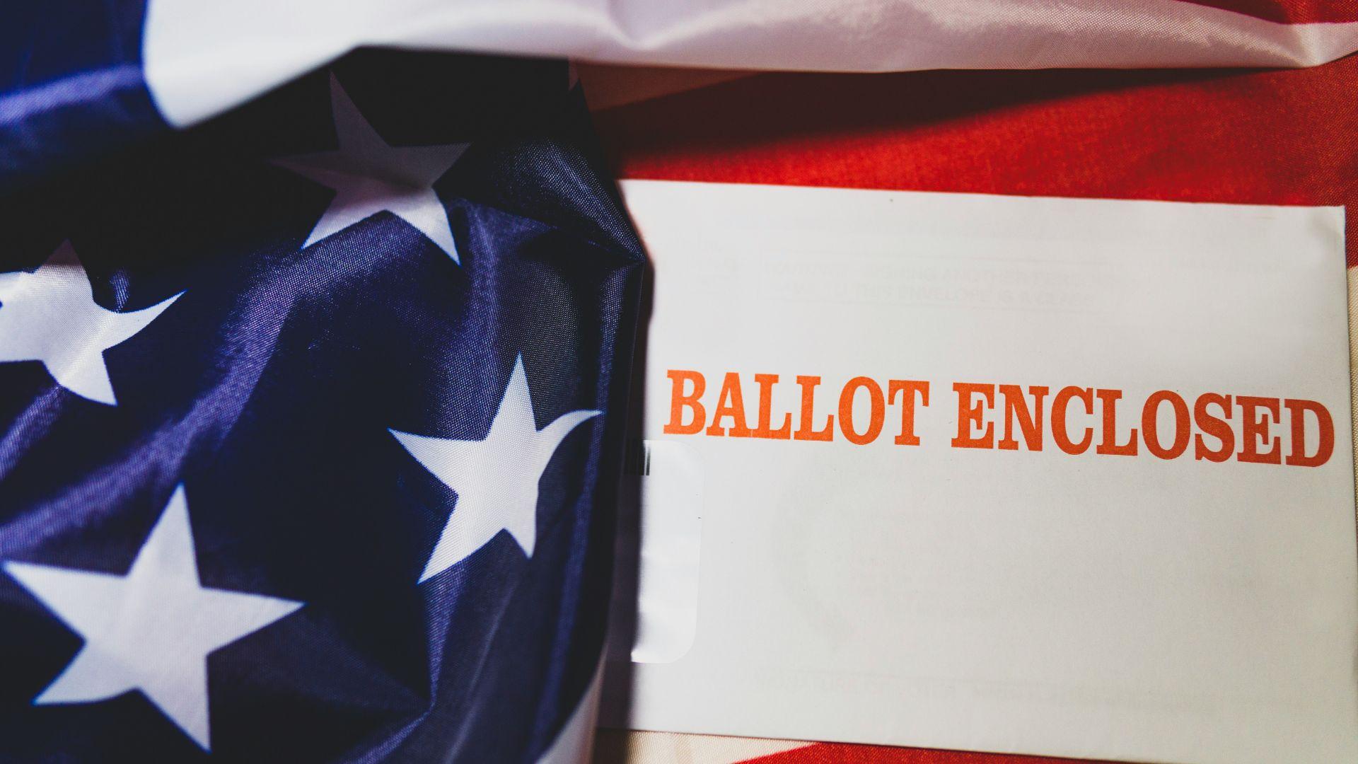 A white ballot envelope next to an American flag.