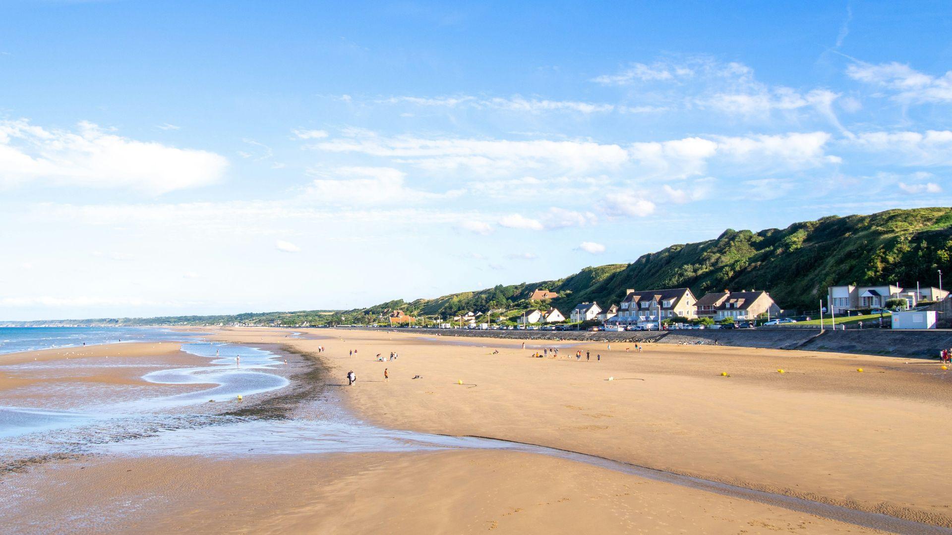 One of the D-Day beaches seen today underneath a blue sky with people walking on the beach.