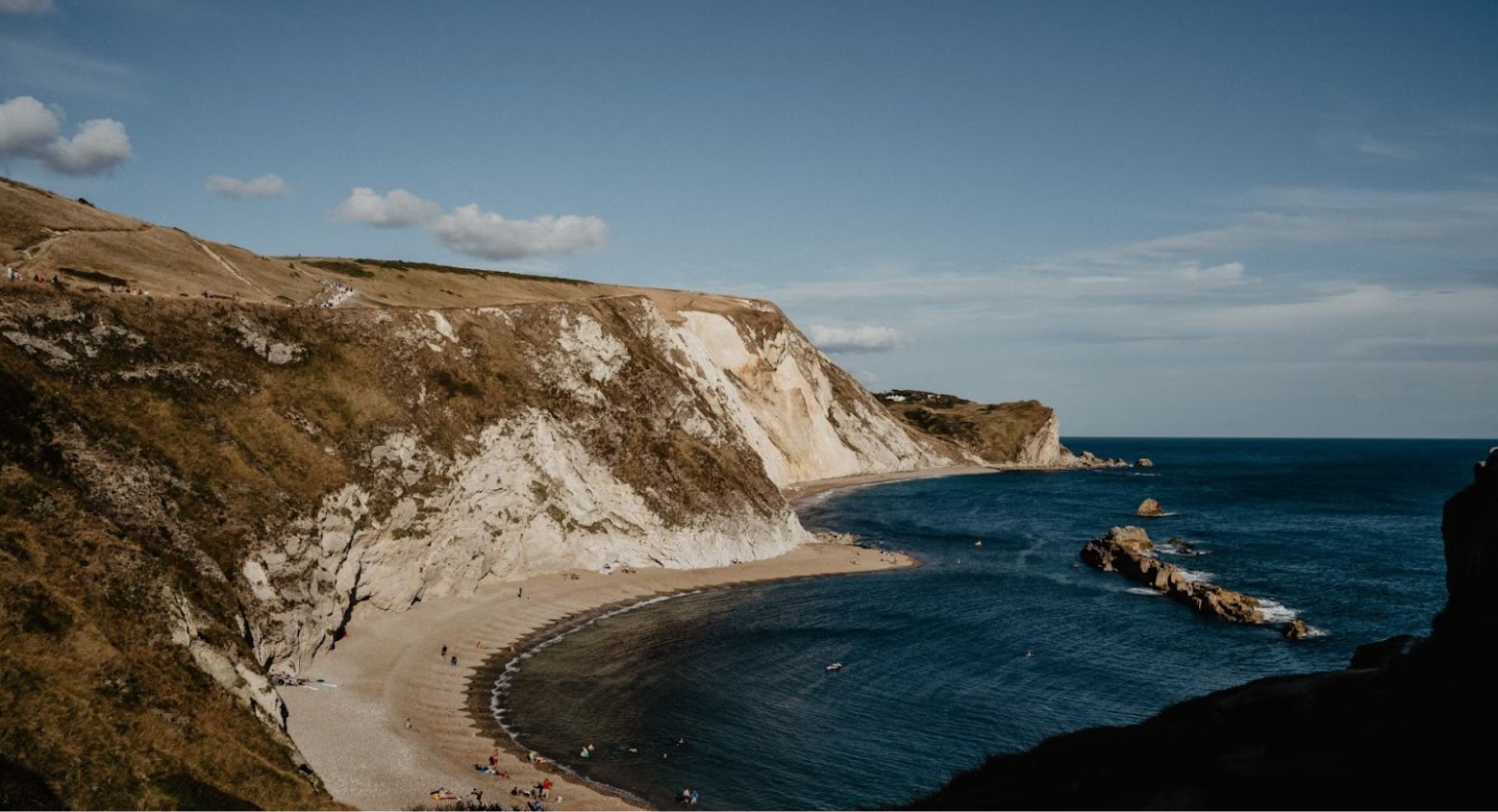 A beach during daytime. The beach is situated next to a cliff. There are some rocks in the sea.
