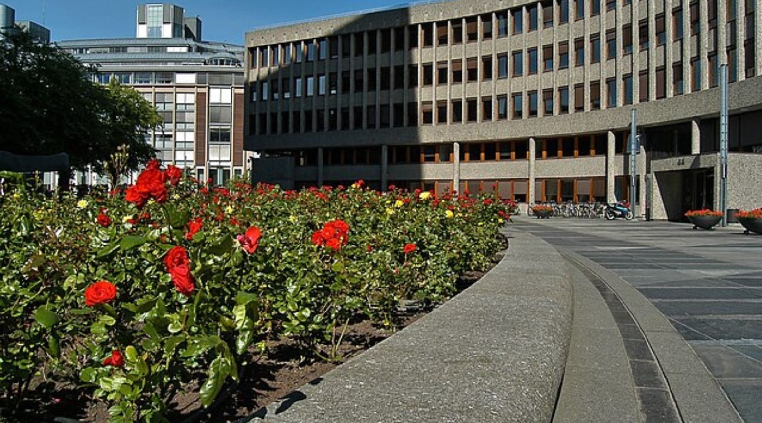 A Government building in Oslo, Norway. The building is curved and there is a garden outside with red flowers and trees planted.