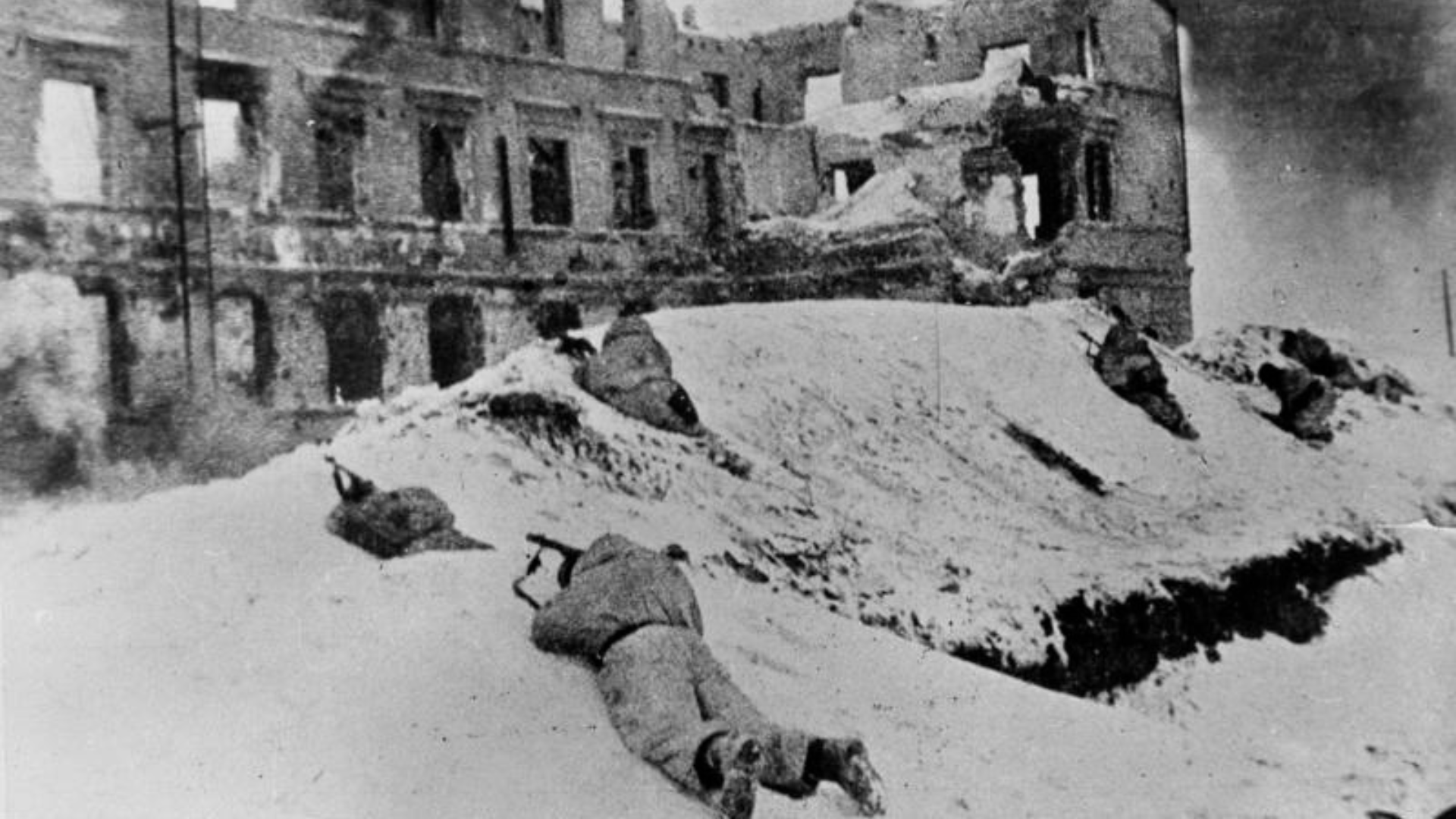 Black-and-whie photo of Soviet soldiers at Stalingrad in a snow embankment near a destroyed building, guns drawn.