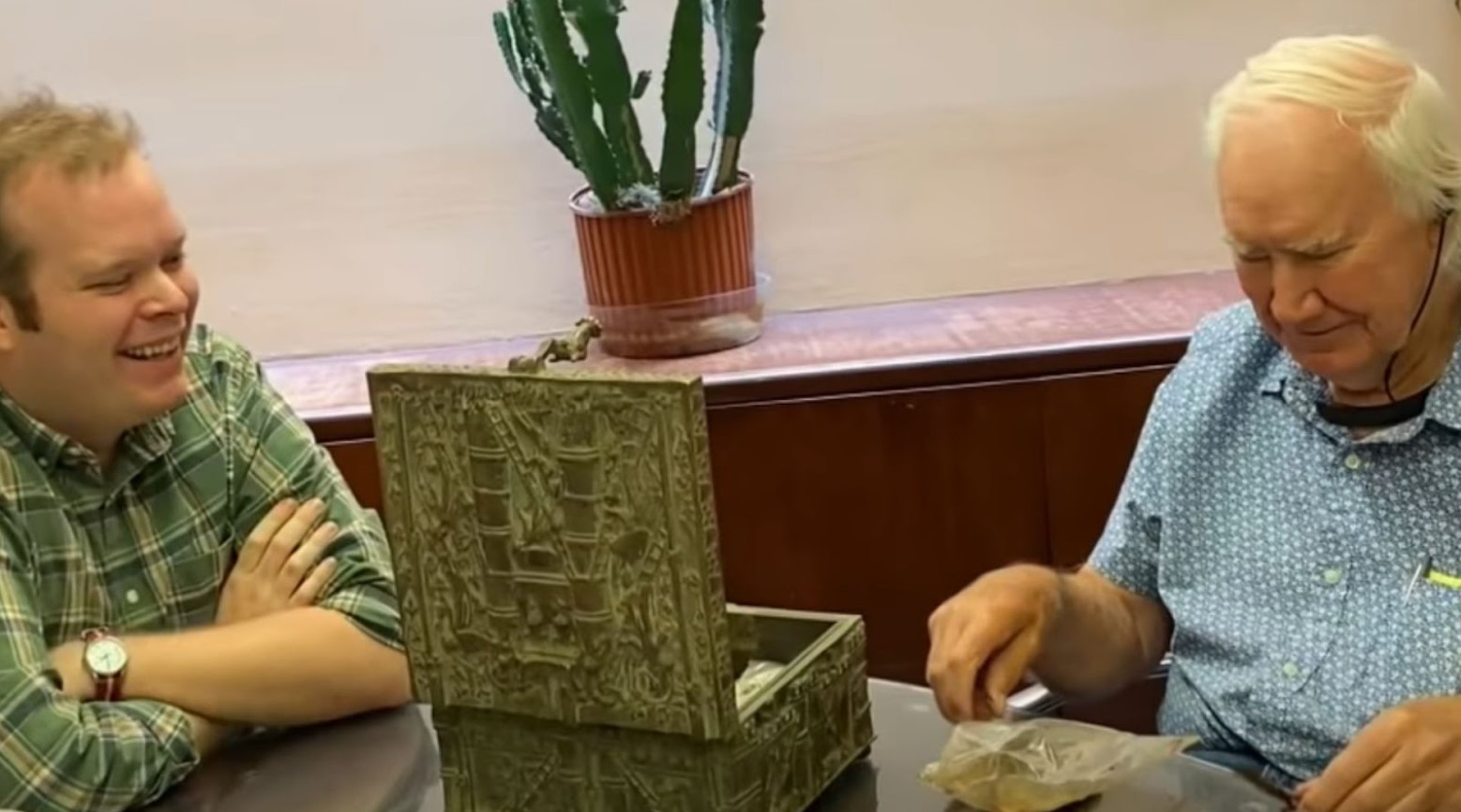 Jack Steuf and Forest Fenn sat at a table with Fenn’s treasure chest in between them. Some of the treasure from inside is in front of Fenn. There is a wooden side unit behind them with a cactus plant on top.