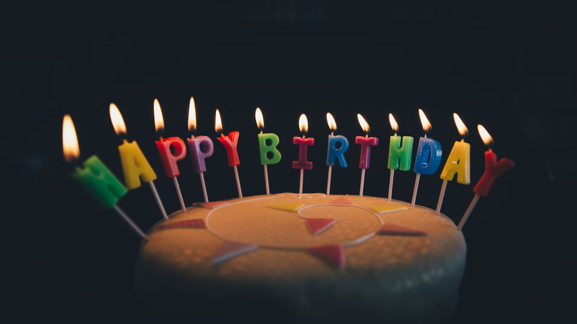 A birthday cake. The cake is decorated in white icing and has candles on that have been lit that spell out "Happy Birthday".