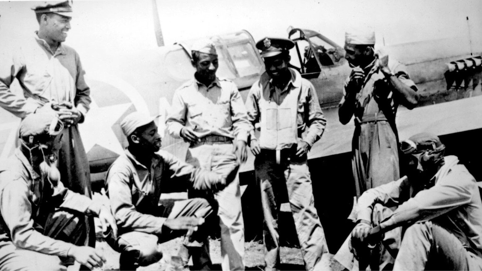 The Tuskegee Airmen sitting and standing around planes while talking.
