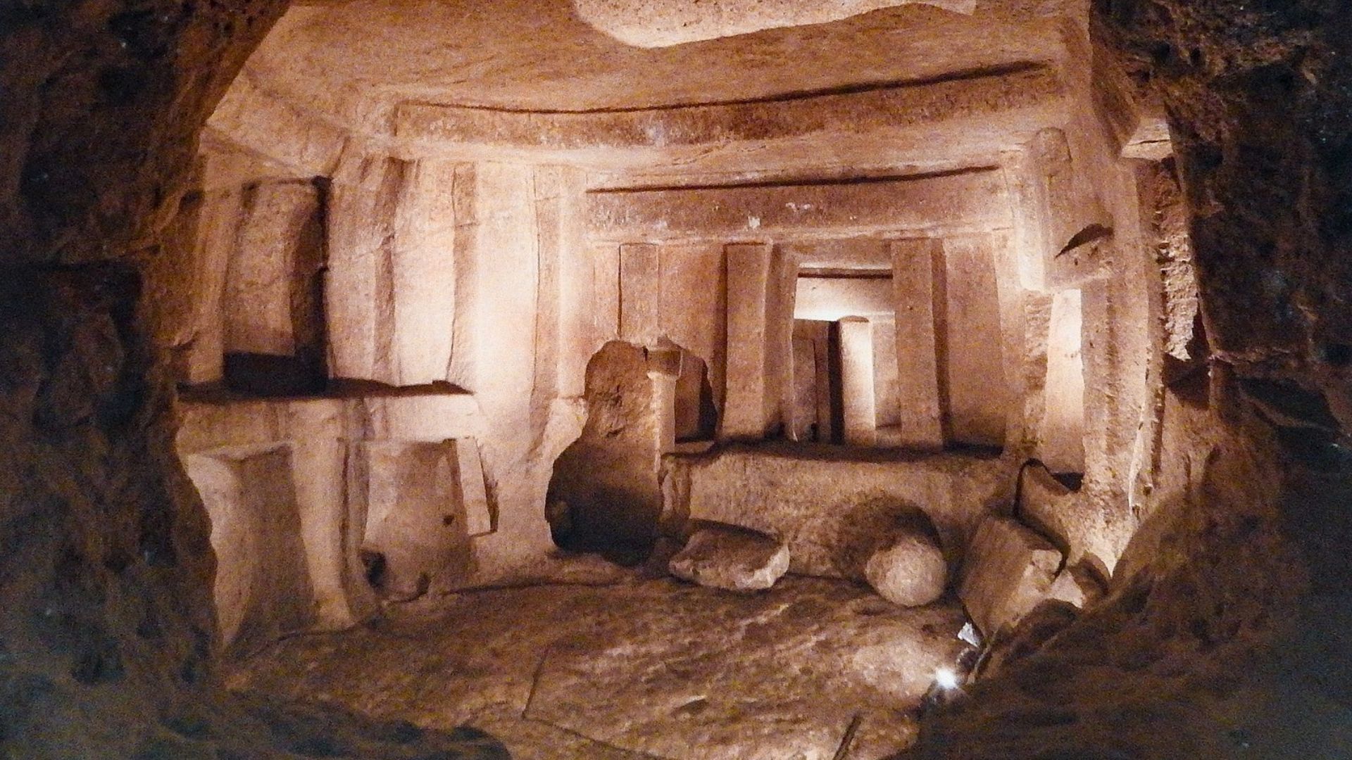 The image shows the interior of the Ħal Saflieni Hypogeum, an underground prehistoric burial site in Malta. Carved entirely out of limestone, the chamber has multiple doorways and alcoves with smooth walls and pillars