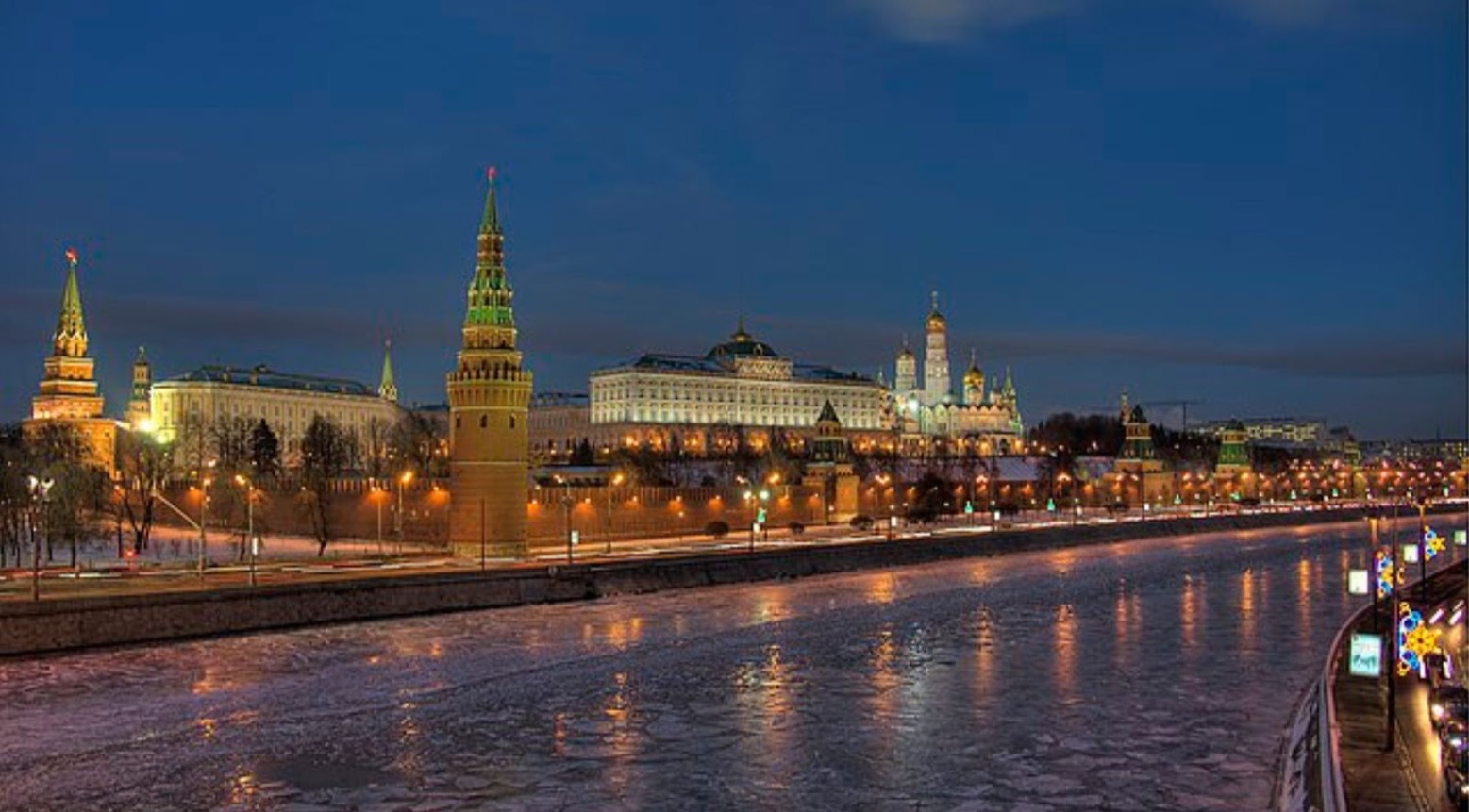 The Moscow Kremlin lit up at night. Lights are running along the outside of the Kremlin, with these lights being reflected into the water.