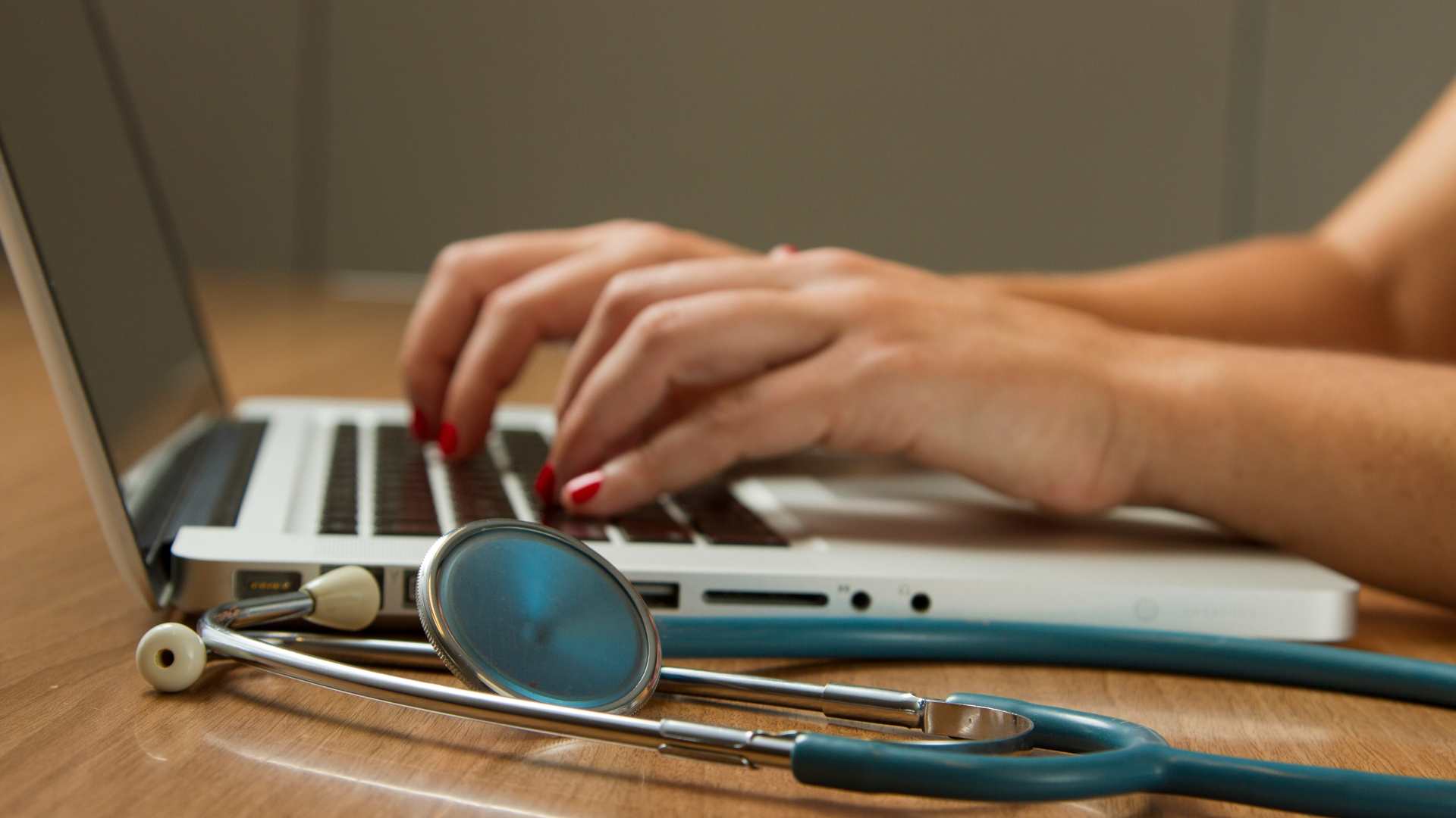 A person types on a laptop while a stethoscope lies next to them.
