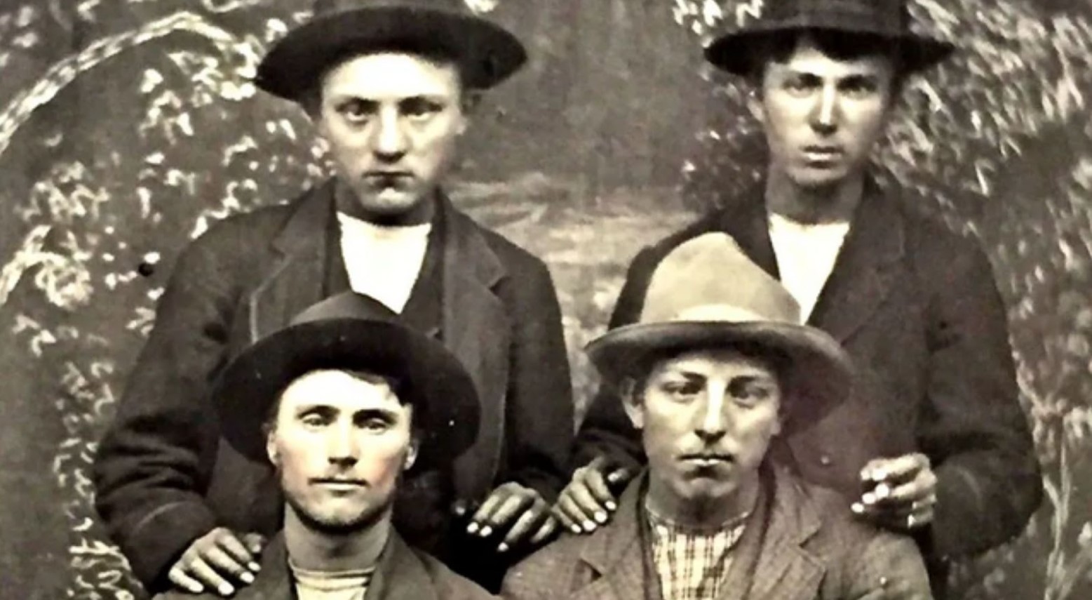 A black and white photo of Jesse James, who is standing next to Emmett Dalton. They are both stood behind Jesse Evans and Frank James. They are all wearing hats and coats.