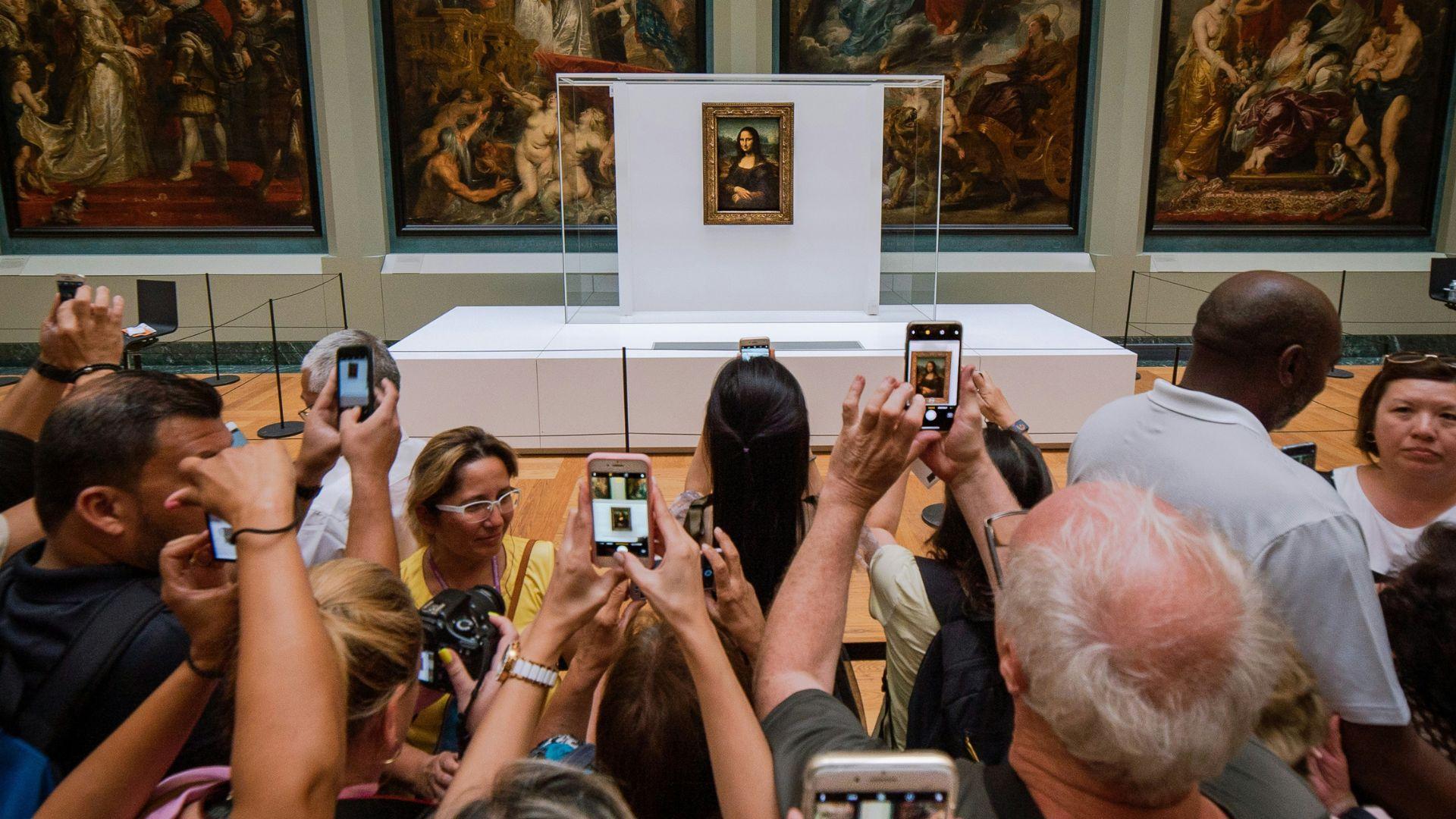 Many people with phones and cameras take photos of the Mona Lisa behind a glass wall in a museum.