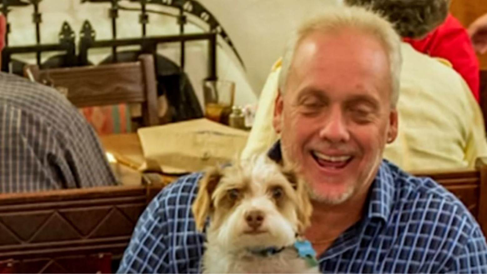 Randy Bilyeu holding his dog Leo. Bileu is wearing a blue chequered shirt and is smiling but has his eyes closed. He appears to be in a restaurant, with people sitting behind him eating food.