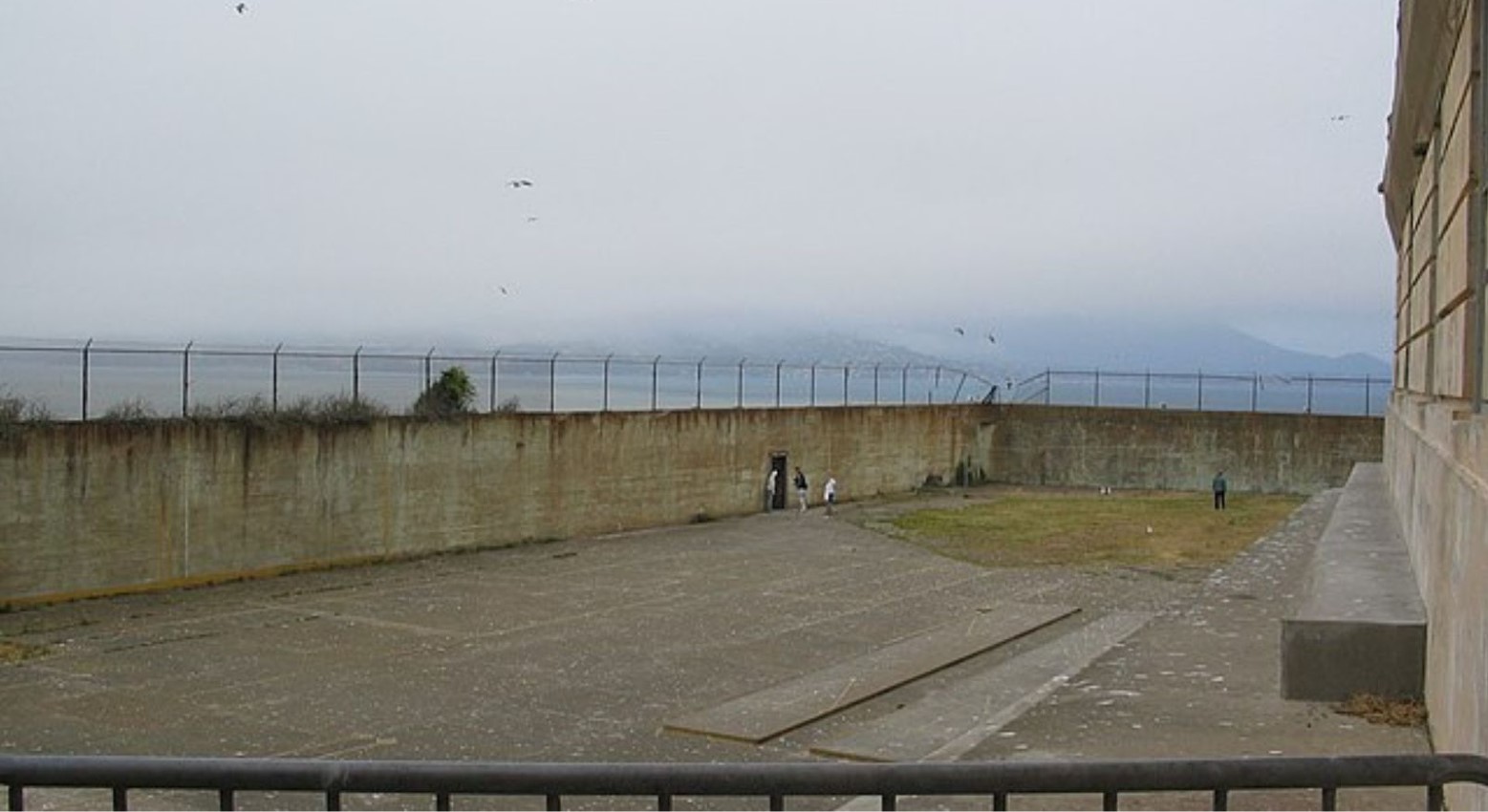 The Alcatraz prison recreation yard. The yard is made of concrete and has a high wall surrounding it so no one can get out. There are some hills in the distance covered by clouds.
