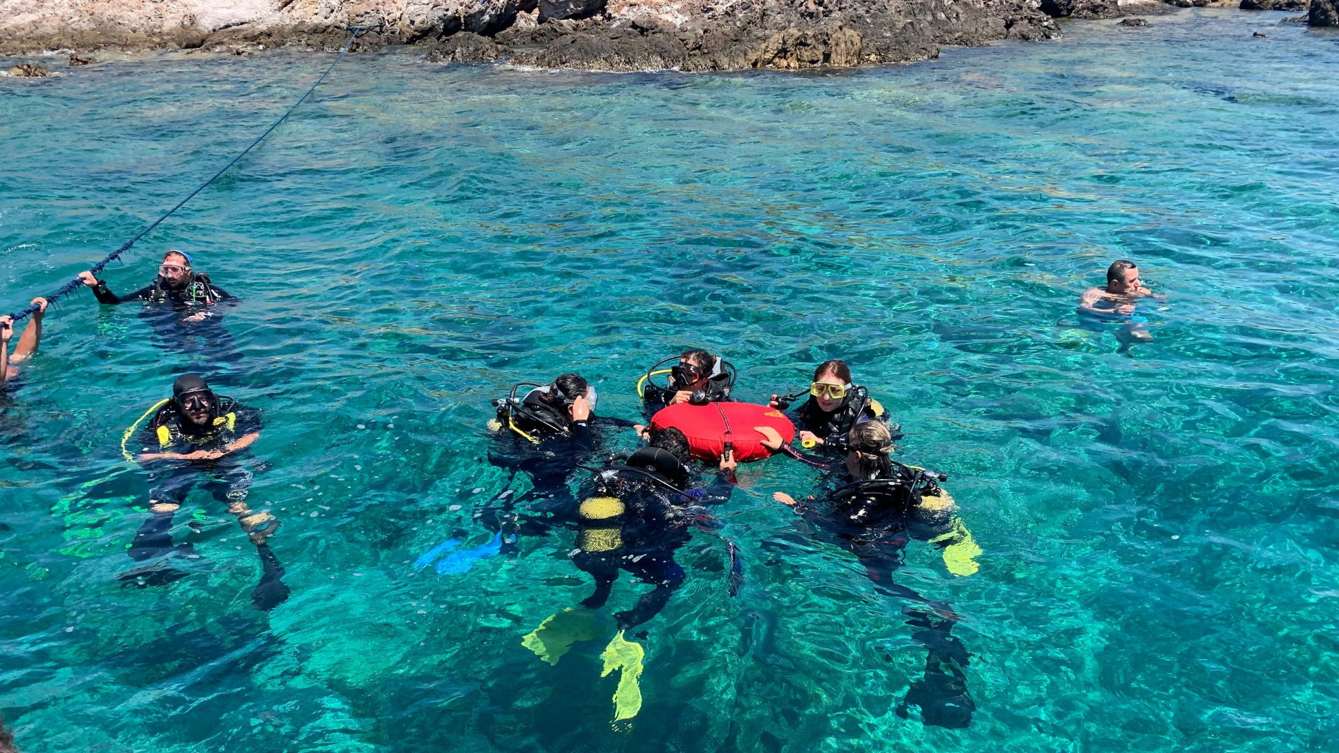 A group of scuba divers is floating on the surface of crystal-clear turquoise water near rocky shores. They are equipped with full scuba gear, including yellow fins, black wetsuits, and dive masks. Some divers are holding onto a line extending into the water, while others seem to be in a briefing or instruction session with a dive leader who is holding a red buoy