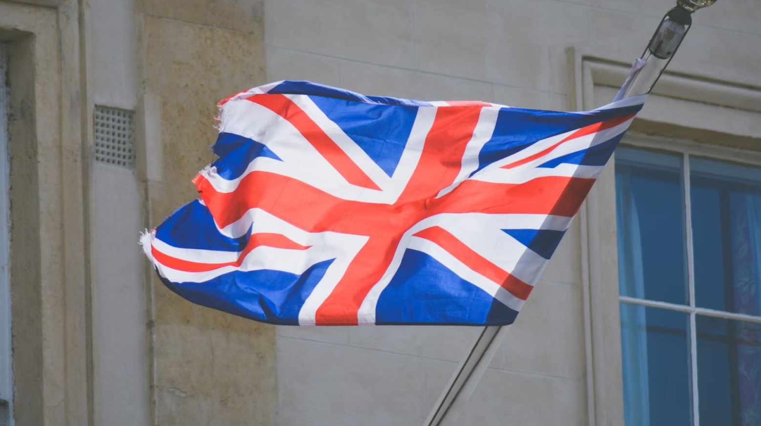 The UK flag flying on a flag pole that is coming out of the side of a building. The UK flag consists of a red cross with a white background, four diagonal red lines against a white background, and all of these against a blue background.
