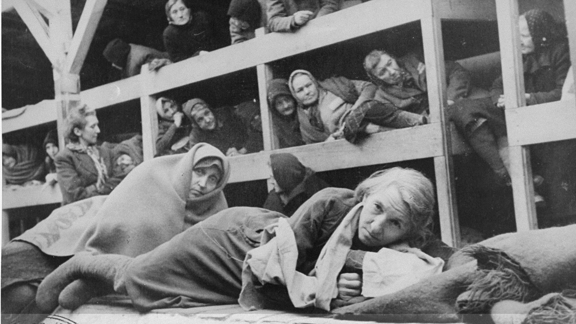 Black-and-white photo of numerous women lying in crowded barracks beds or on the ground at the Auschwitz concentration camp.
