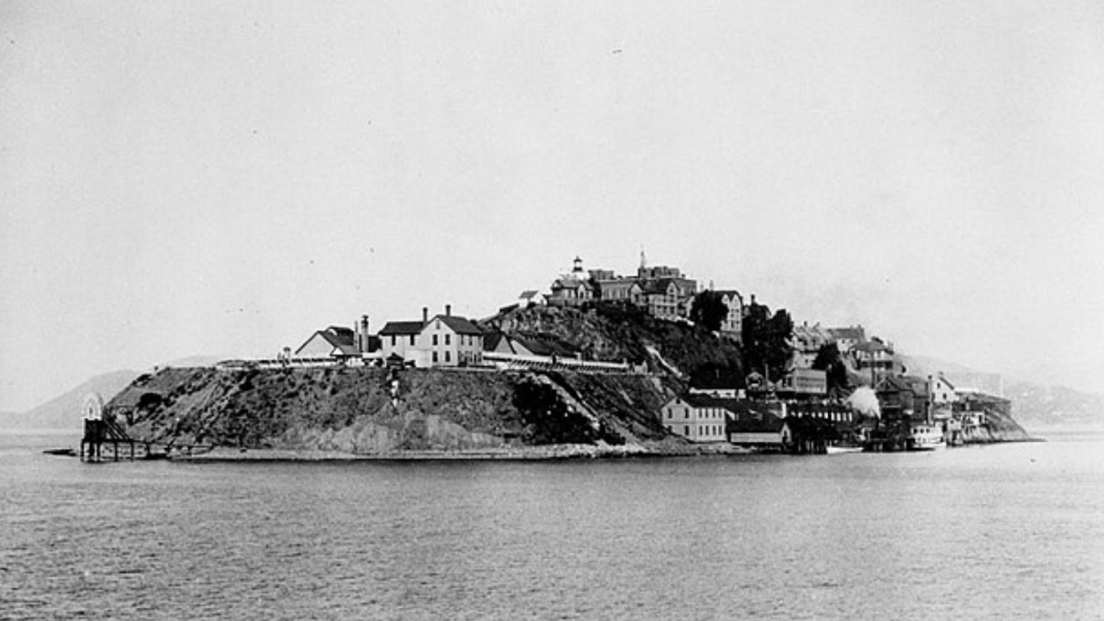 A black and white photo of Alcatraz Island before the prison was built. There is a jetty at the bottom of the island and various houses and other buildings are built along the island.