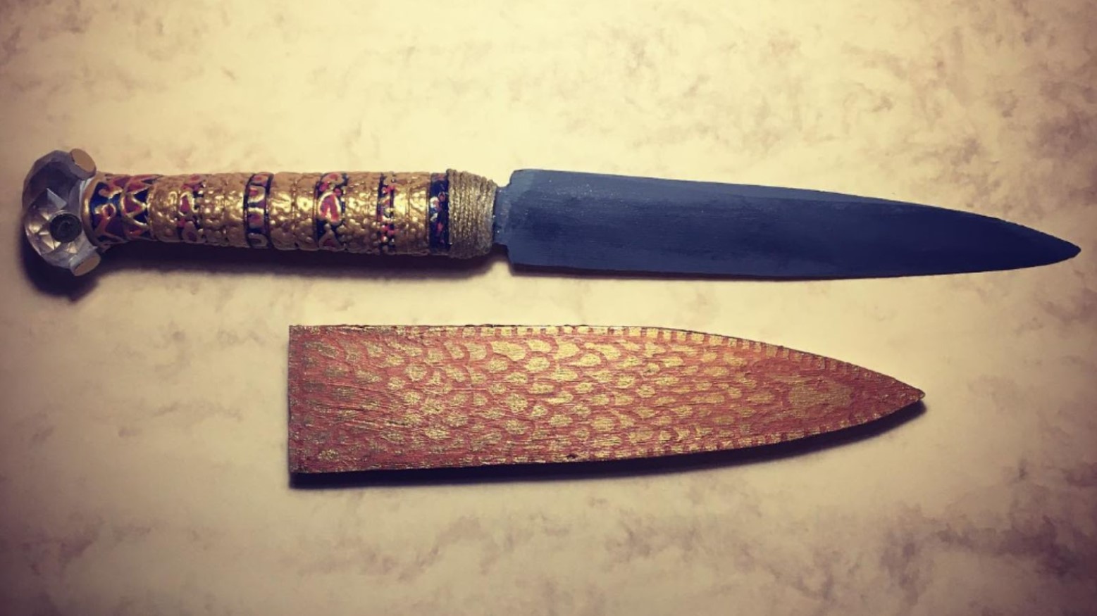 King Tutankhamun’s dagger on a white background. The handle of the dagger is gold with a blue and red pattern and the dagger itself is a dark gret. It also comes with a cover that is gold and red.