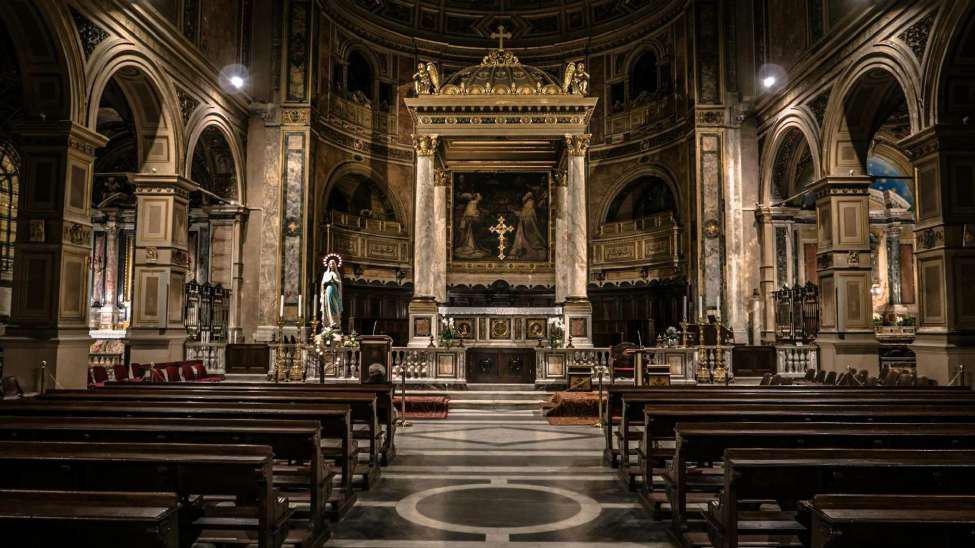 The inside of an empty Church. There are wooden benches on either side of the aisle and at the front of the church is a big cross and a statue. There are paintings throughout on the walls.