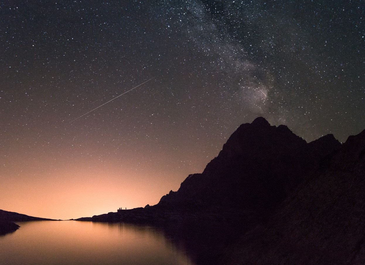 A very starry night sky over mountains and a body of water at night.