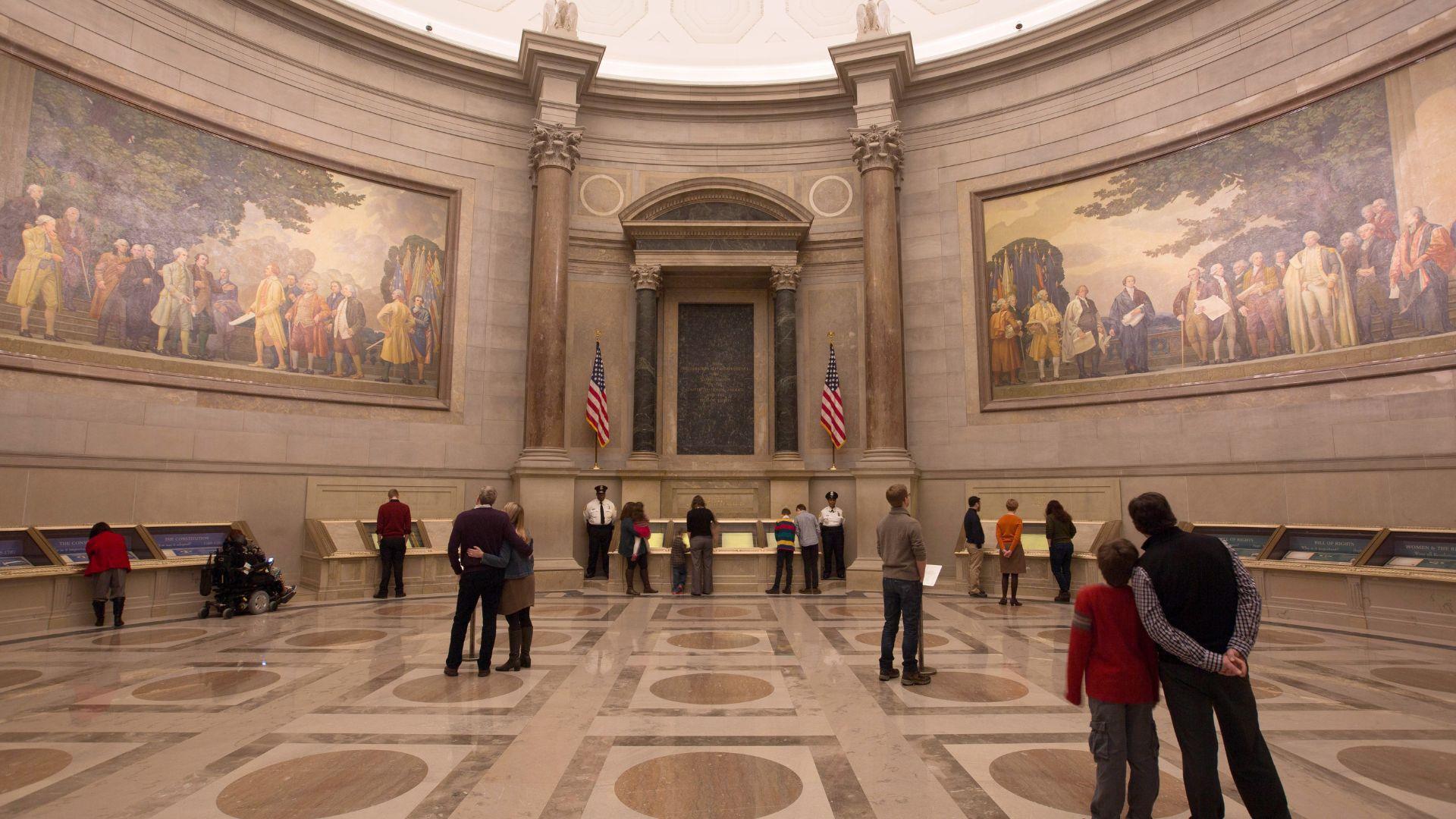 An interior view of the National Archives’ rotunda, with paintings on the wall, as people visit it.