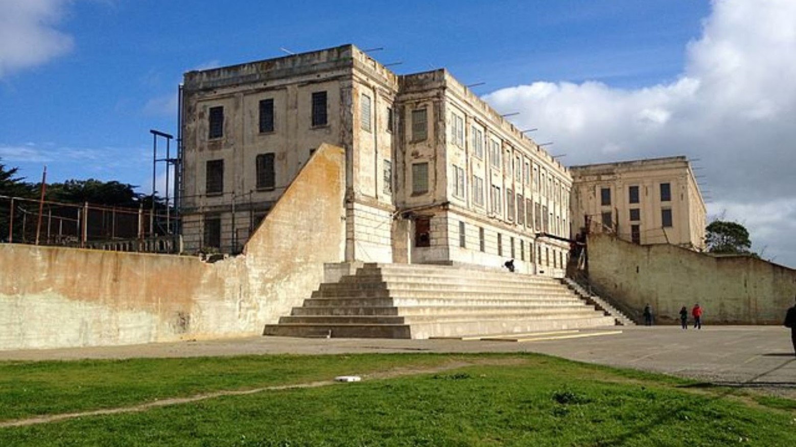 The Alcatraz prison recreation yard. The yard is a mixture of concrete and grass and is surrounded by steps to get down to it and a high wall around it, with the prison in the background.