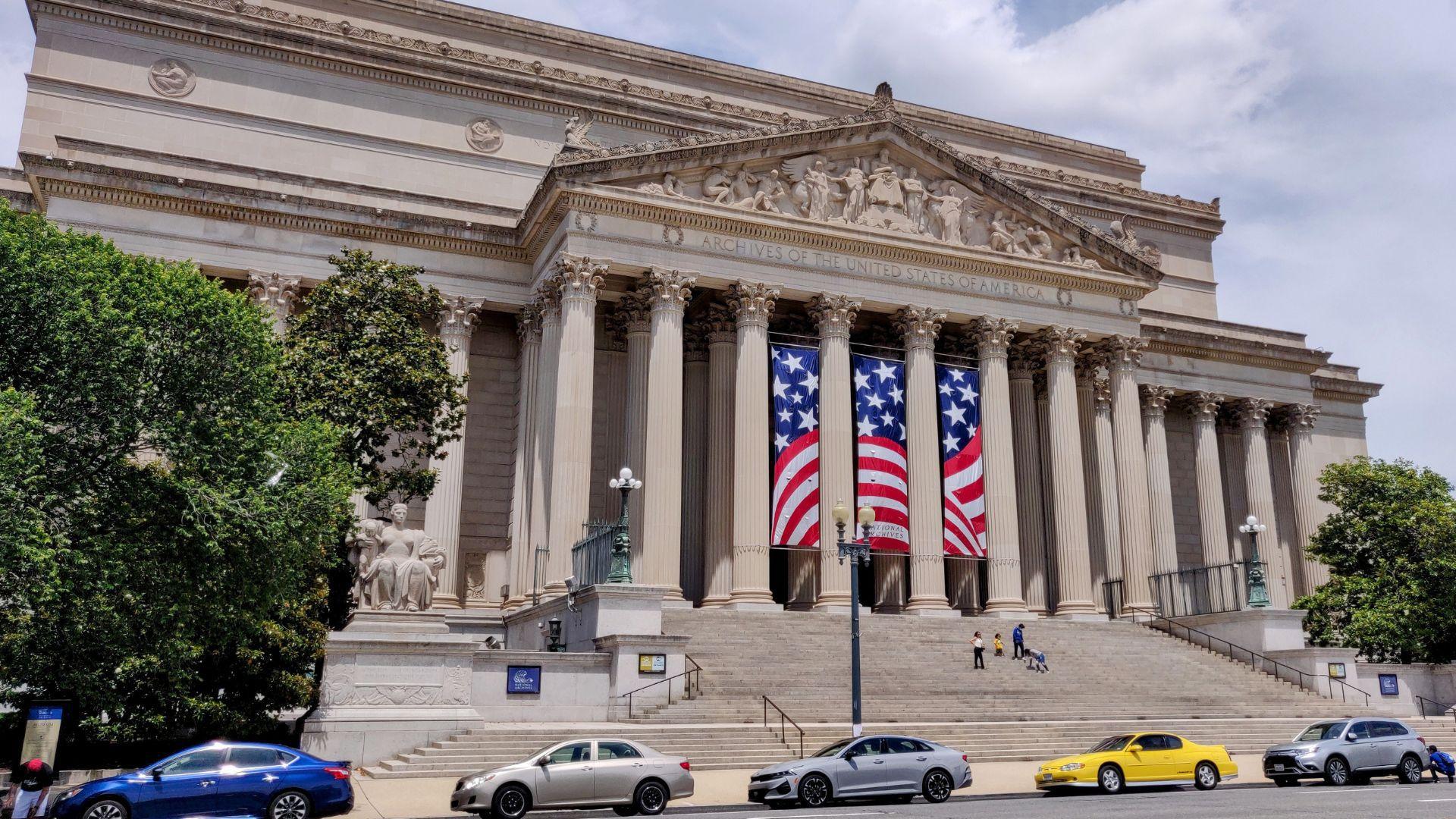 The National Archives building in D.C. with cars in front of it and people walking up its steps.