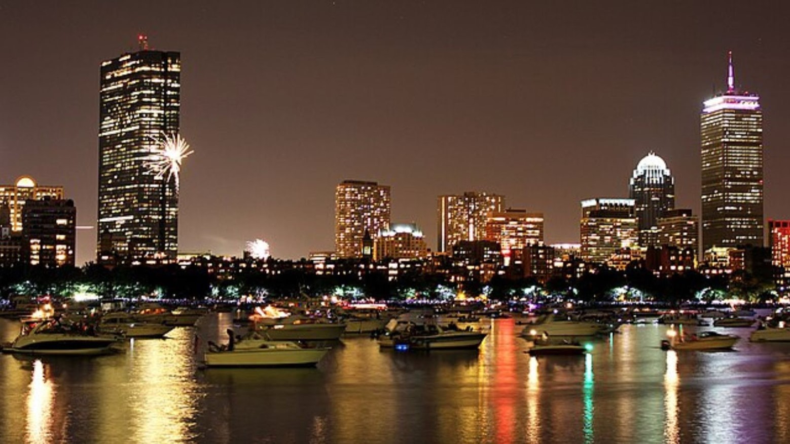 The Boston Harbour at night. There are boats in the harbor and buildings in the background that have been lit up, with the lights reflecting in the water.