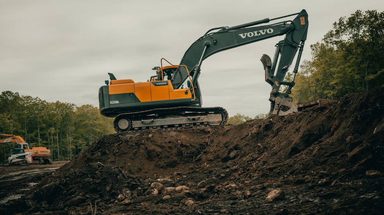 A digger digging up land at an excavation site. It is on top of some mud and trees surrounding the site. The digger is yellow and black, is on caterpillar tracks, and has ‘Volvo’ on the arm.