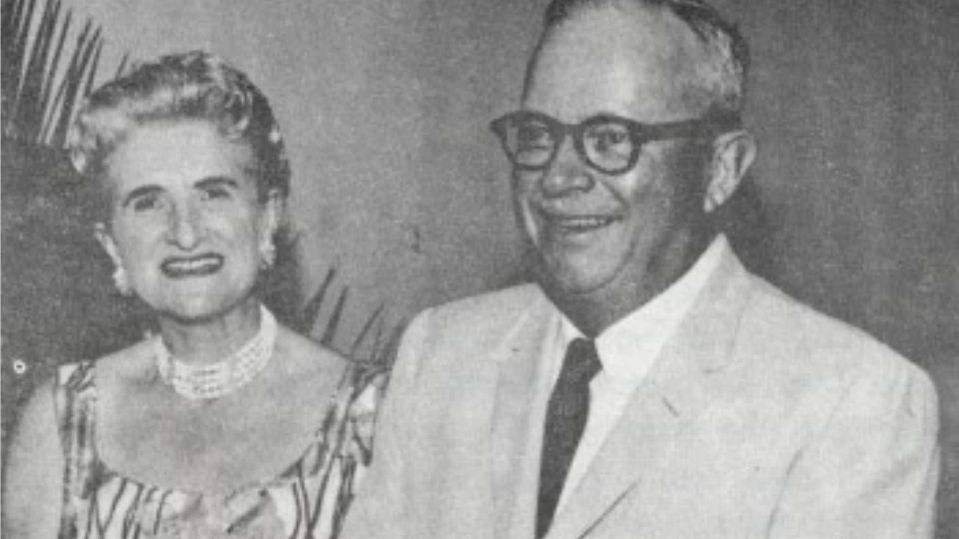 A black and white photograph of an elderly couple posing for the camera. The woman, on the left, is wearing a dress with a wide-striped pattern and a pearl necklace. She has styled hair and is smiling. The man, on the right, wears glasses, a white shirt, and a light-colored suit. He is also smiling