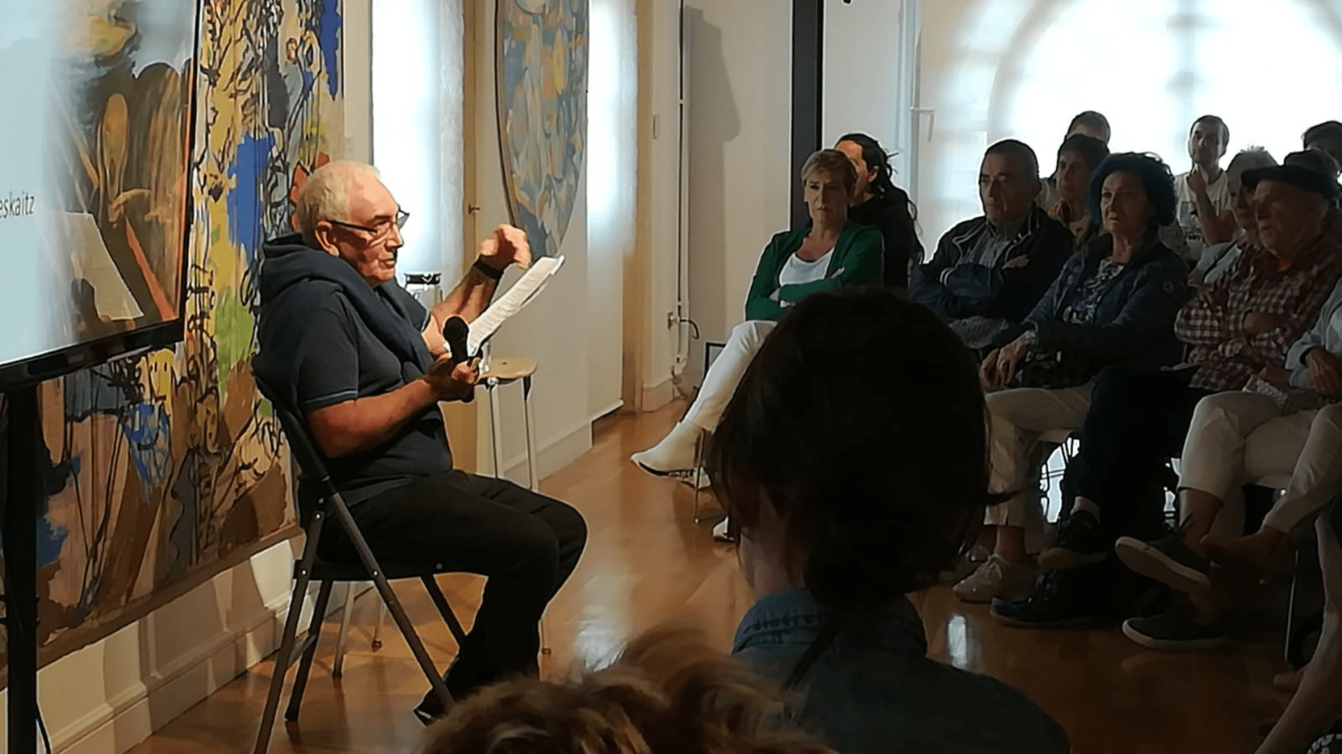 A Basque literary and cultural gathering under the organization “Hitzez eta Ahotsez.” People sit in a semi-circle around an elderly man who is sitting and speaking.