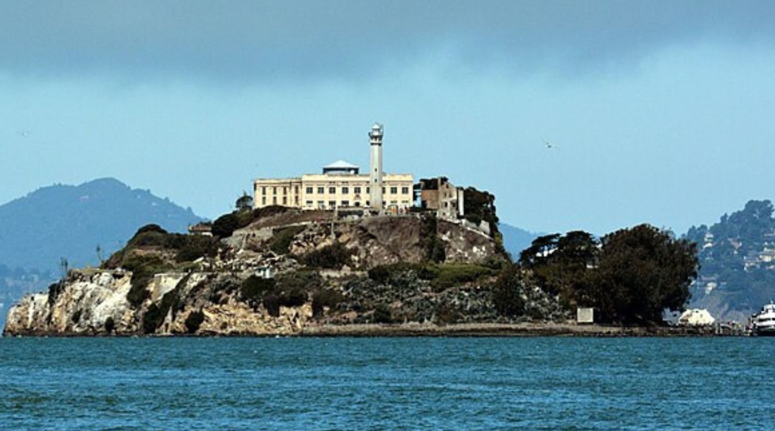 Alcatraz Island. The prison is at the top of the island and some hills can be seen in the distance behind it. The island is surrounded by blue sea and a lighthouse is in front of the prison.