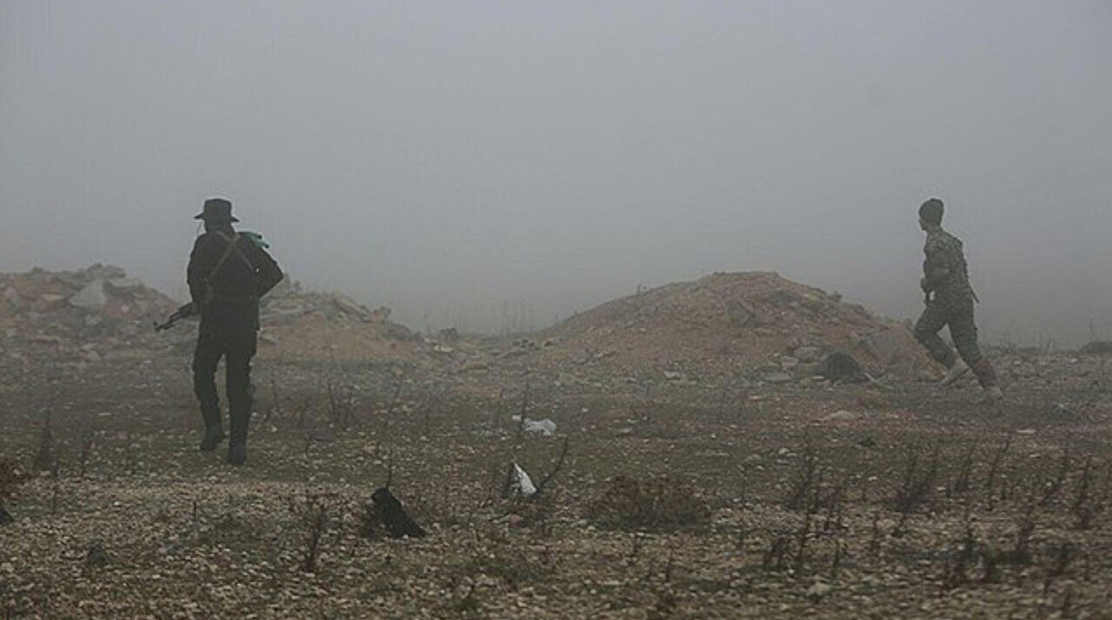 An image from the Syrian War. Two soldiers are walking and the land they are walking on is mostly rubble. Some parts are flat and others are mounds of rubble.