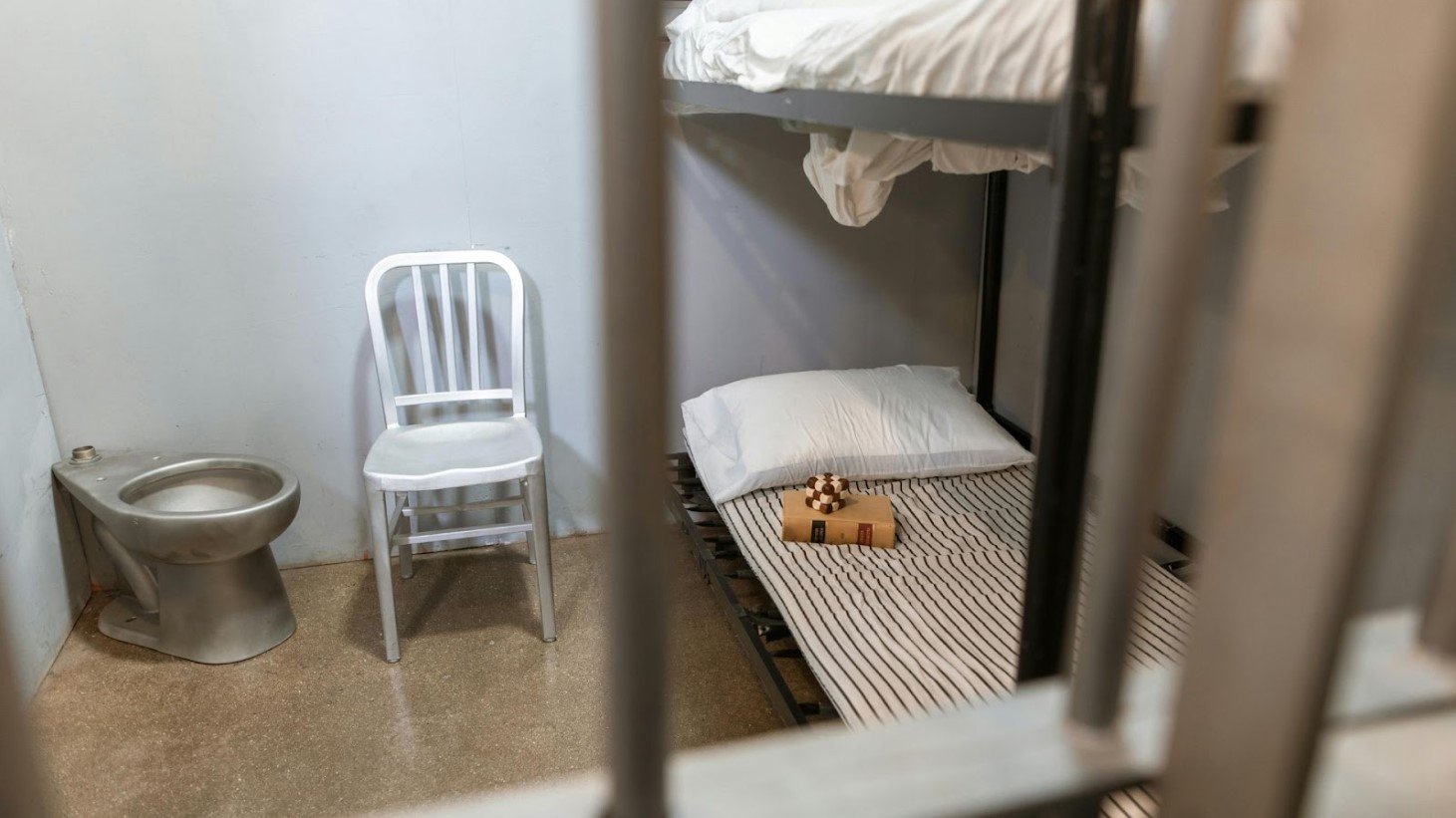 View of a jail cell interior with a stainless steel toilet, white plastic chair, and a bunk bed with a white pillow and a chess set on the lower bed