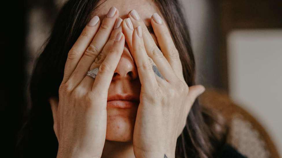 A woman with her head in her hands. She has silver rings on her ring finger and a tattoo on her opposing finger.