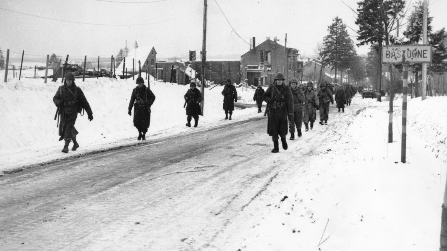 Two rows of soldiers of the 101st Airborne Division walking out of a town in the snow during the Battle of the Bulge in World War II.
