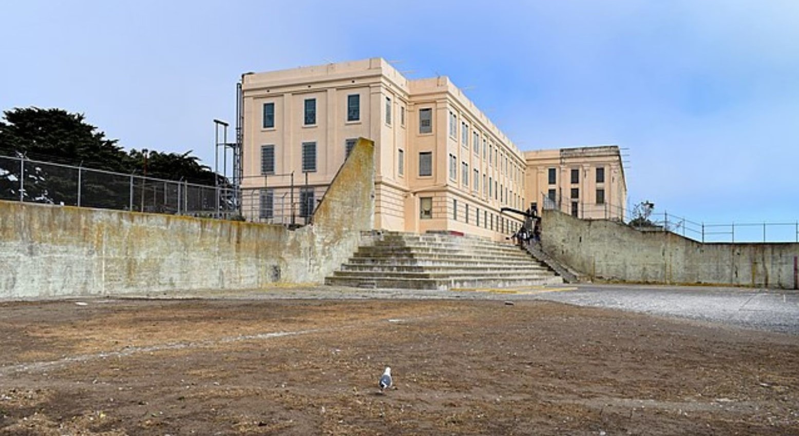 Alcatraz prison in the recreation yard. The yard is mostly dirt and has some steps leading up to the prison behind it. There is a barrier of high walls all around the yard.