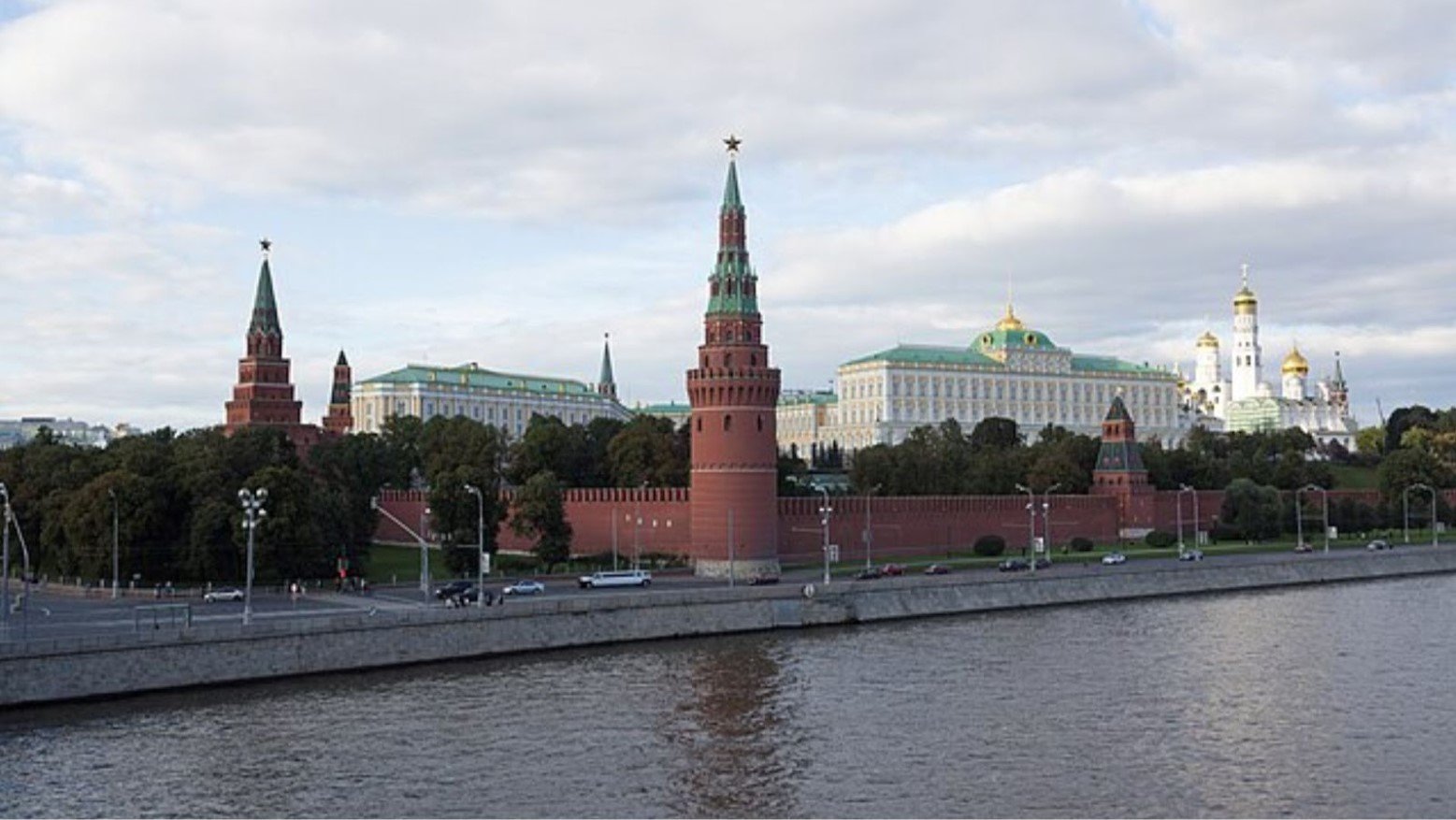 The Moscow Kremlin. The walls are red with turrets that have a green top. The building is white, with a green roof and gold on top. It is positioned next to the water.