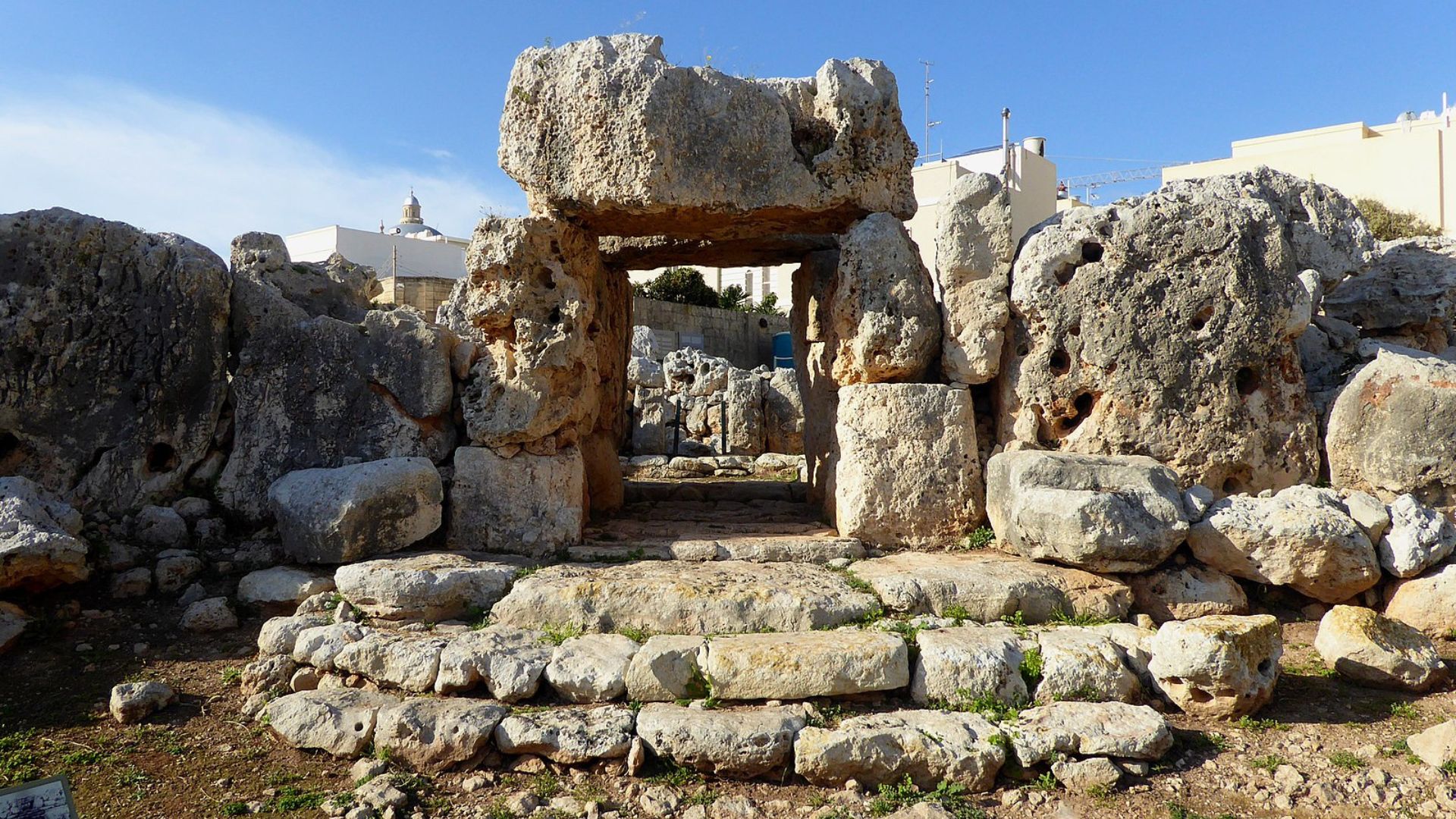 A photograph showing a megalithic structure with a large stone doorway composed of three huge limestone blocks – two vertical standing stones capped by a massive horizontal lintel. The structure is part of a larger pile of boulders and stone ruins