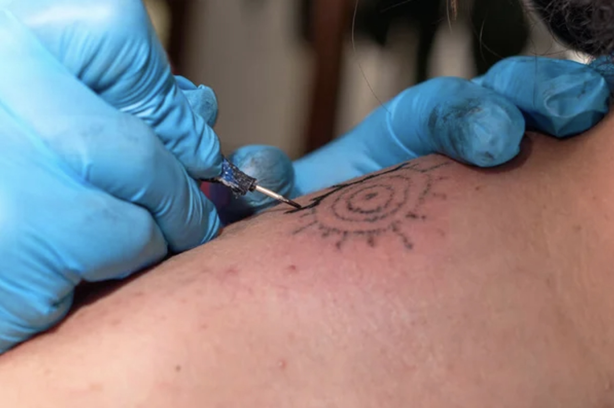Two blue-gloved hands administering a stick-and-poke tattoo on someone’s skin