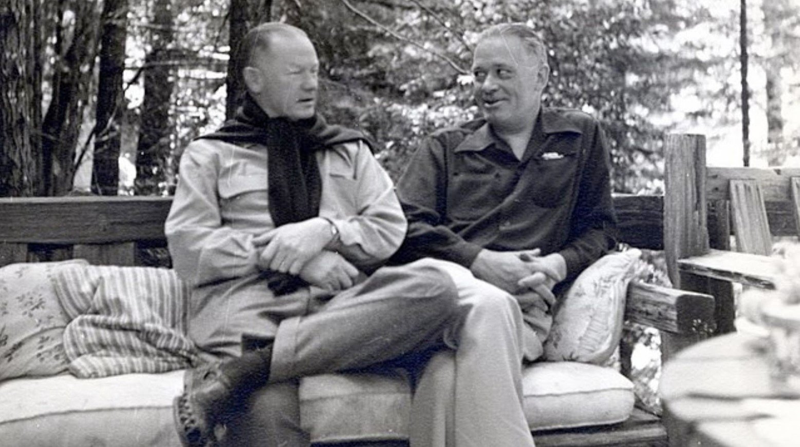 This is a black-and-white photo of two men at the Bohemian Grove. They are sitting on a chair with cushions, and trees are in the background.