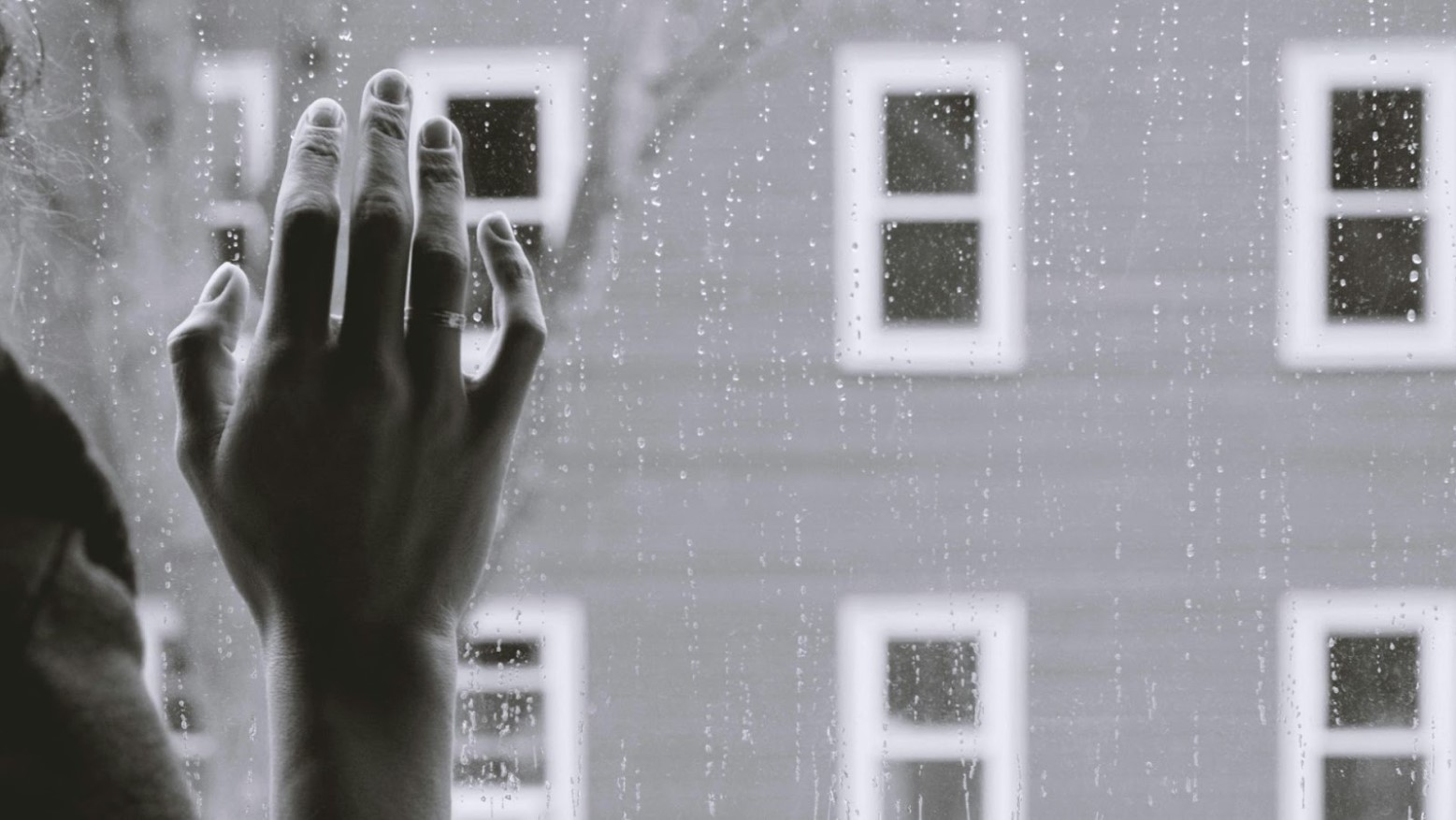 A grayscale photo of a person’s hand on a rainy window.