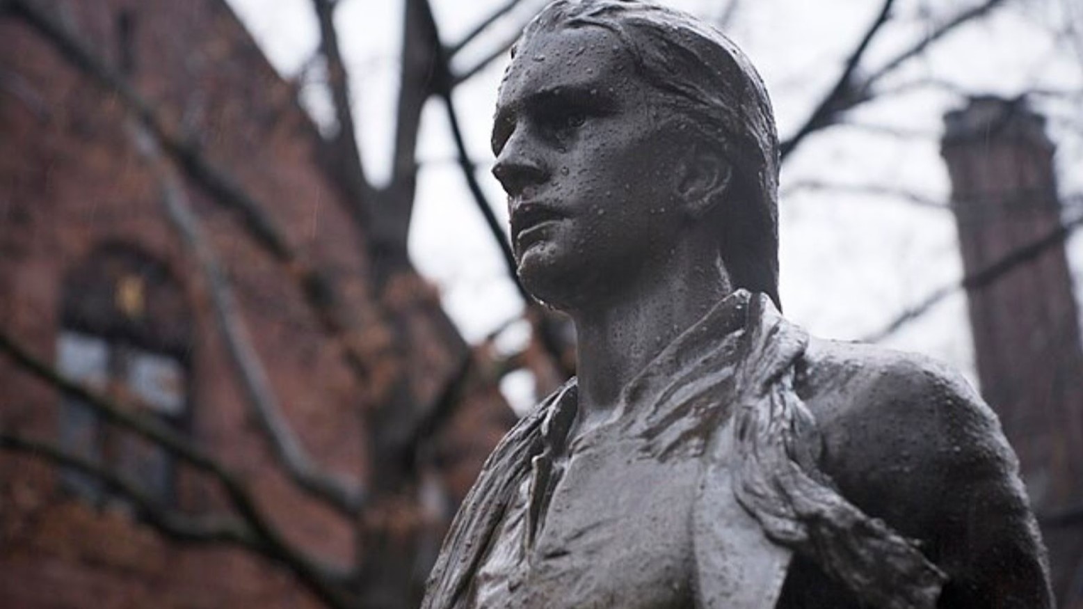 A statue of Nathan Hale. The statue is grey and is in front of a red-brick building.