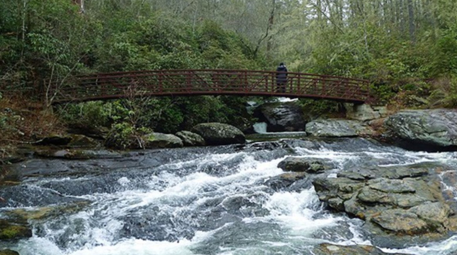 The Norton Creek Trail. There is a red bridge over a river with rocks and a person standing on the bridge.