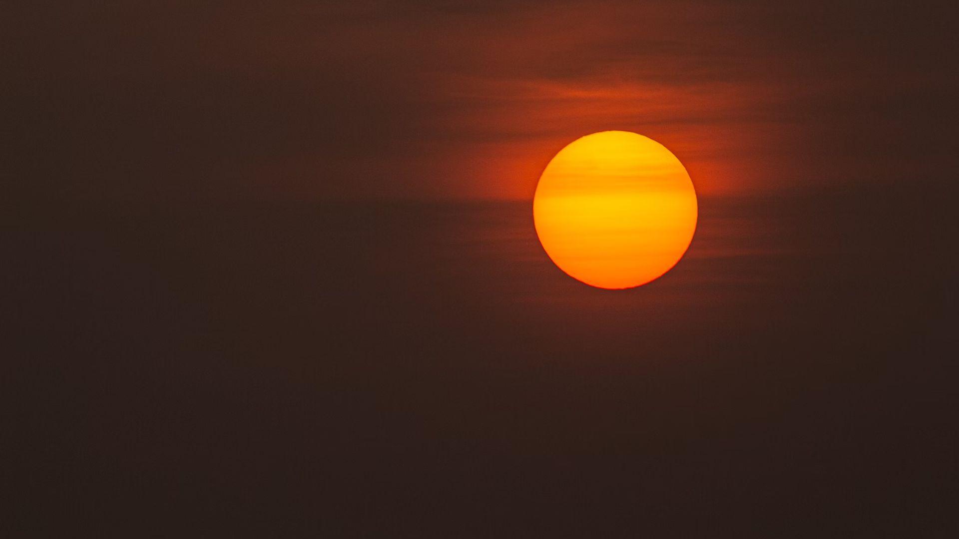 The sun is setting in the dark sky. The sun is orange, and there is a slight haze over it.