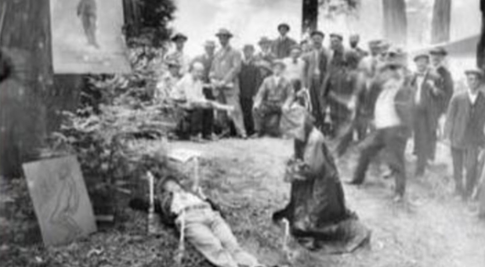 This is a black-and-white photo of a ritual in Bohemian Grove in 1915. A man is lying on the ground, and someone in a hood is kneeling next to him. A crowd of people is watching.