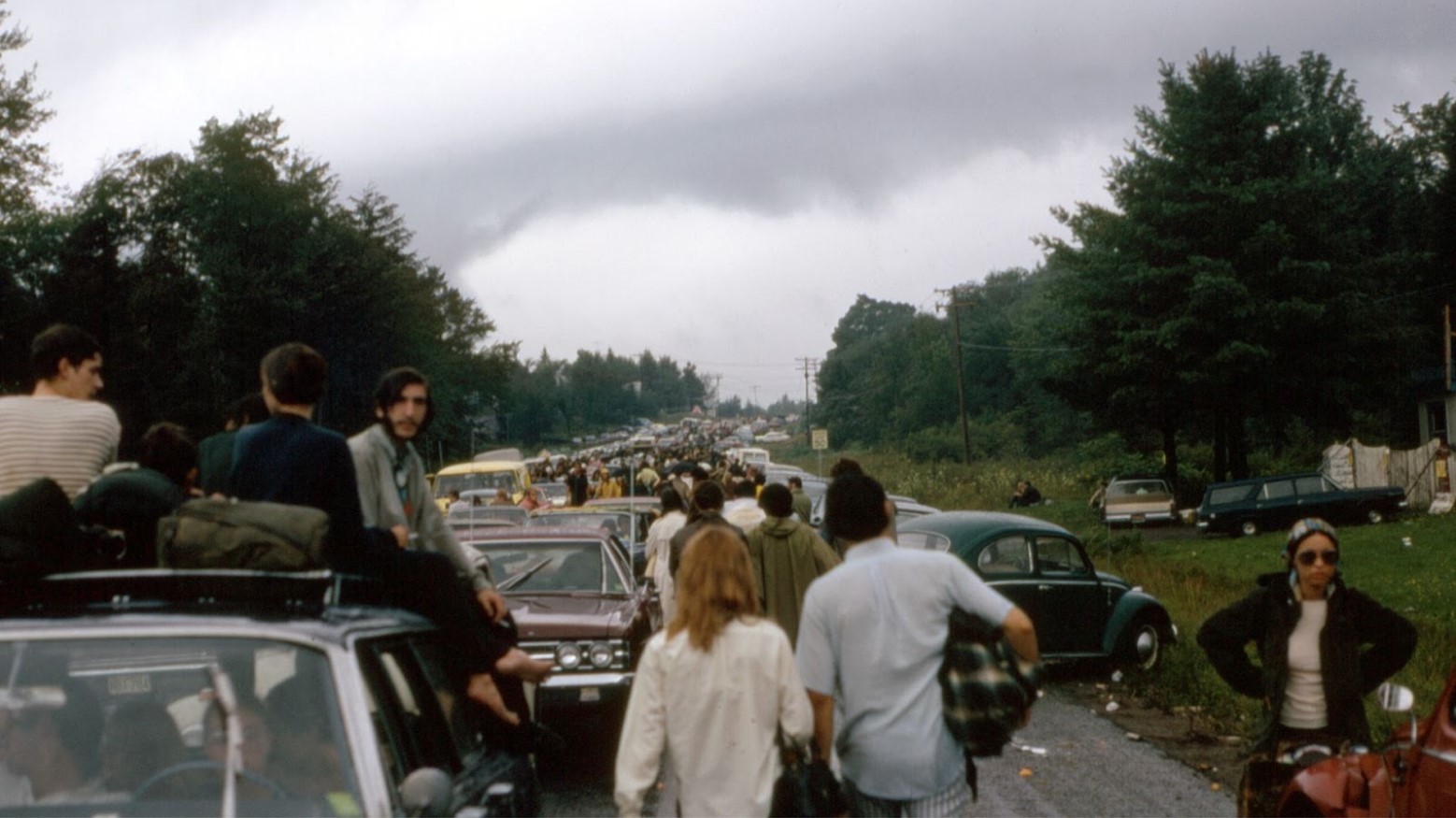 Many people walking, riding on top of cars, and driving cars on the way to Woodstock in 1969.