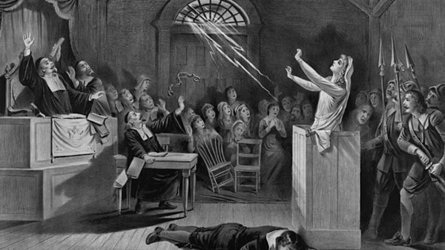 A black and white drawing of the Salem witch trials. The ‘witch’ is in the dock, holding her arms up as if she’s making a spell. There is a man on the floor in front of her. The judge and everyone in the room are in shock. Some are hiding behind chairs.