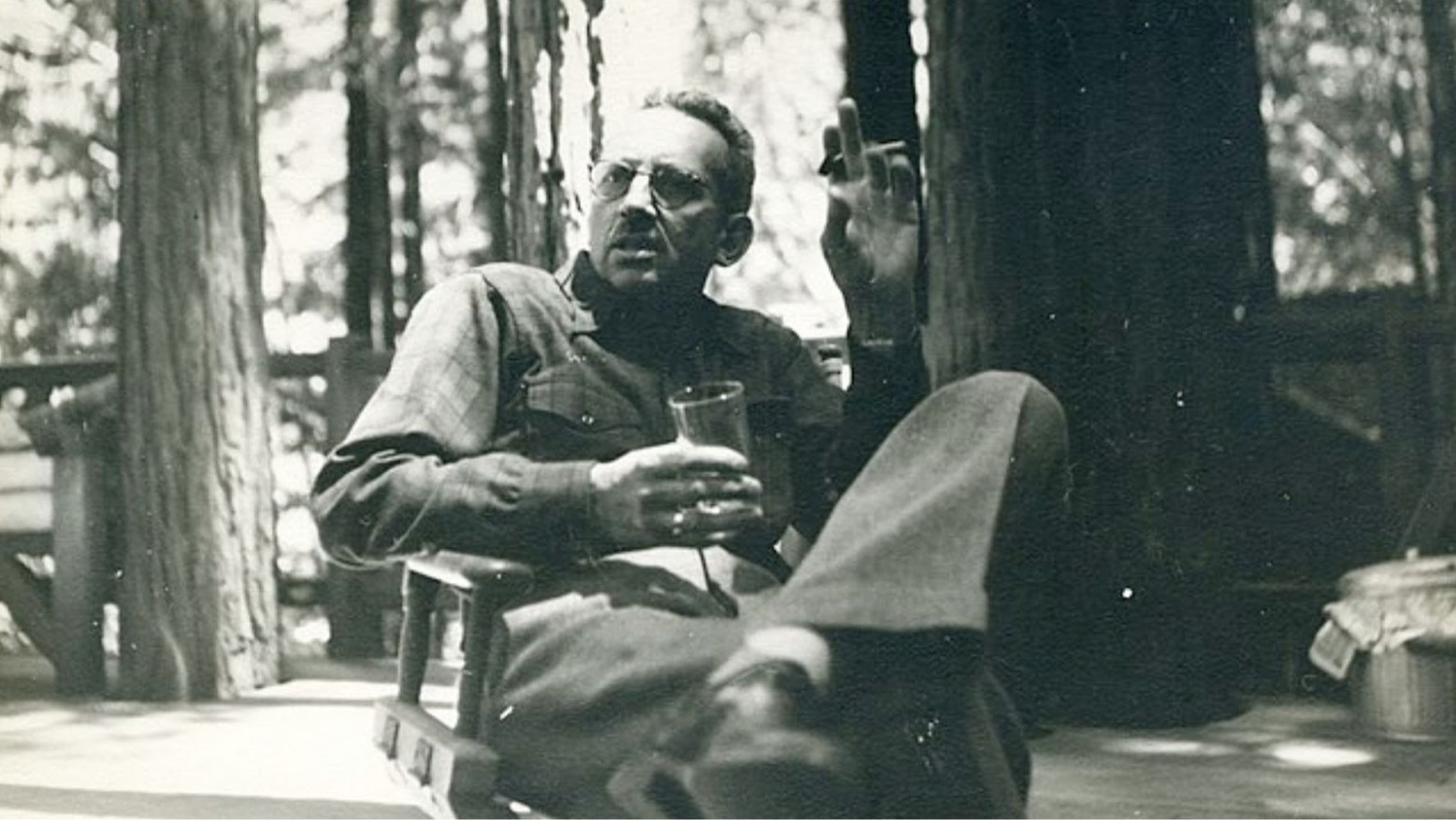 This is a black-and-white photo of a man sitting in a chair at Bohemian Grove. He has a glass in one hand and a cigarette in the other. Trees are in the background.