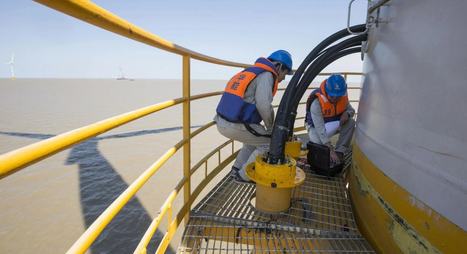 Two men standing on a platform in the middle of the sea that is pumping water. They are wearing blue and orange life support vests and blue hard hats.