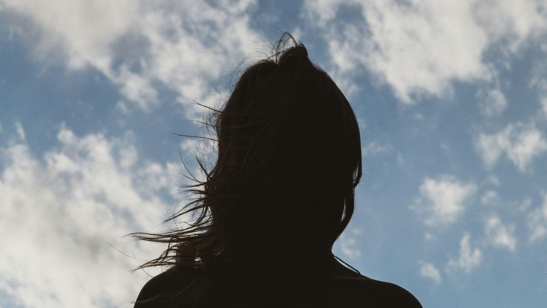 The silhouette of a woman seen outside in the daytime underneath a cloudy blue sky.